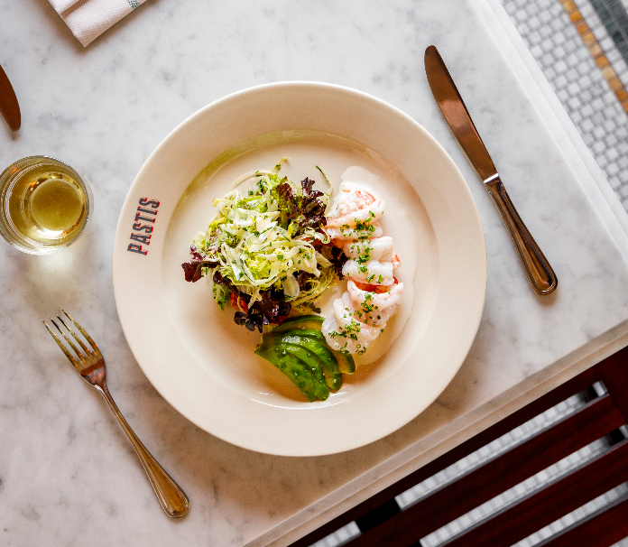 An overhead view of a shrimp and avocado salad on a Pastis restaurant plate, served with white wine on a marble table.