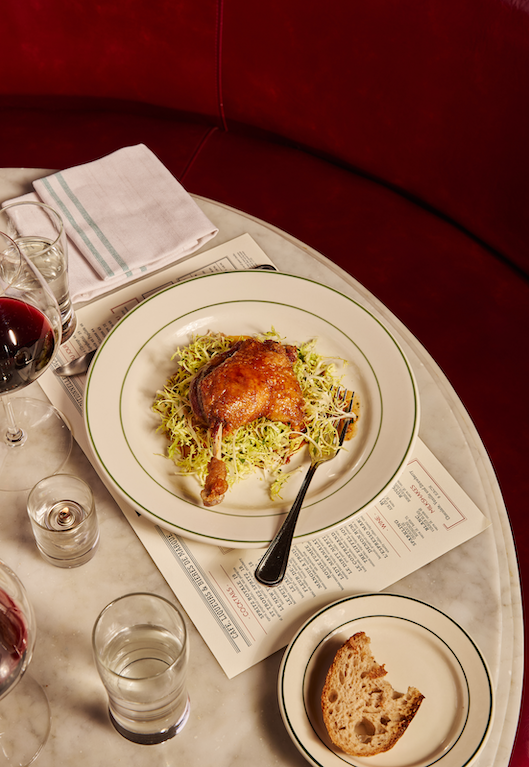 Crispy duck confit on frisée salad, served on a green-rimmed plate atop a printed menu, with red wine, water glasses, a slice of bread, and a napkin on a marble table beside a red velvet banquette.